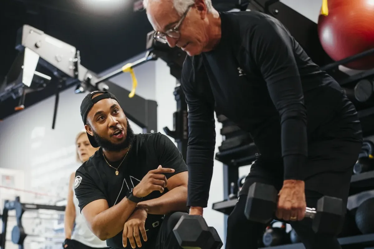 A coach kneels beside an older member mid dumbbell exercise, talking him through the movement at an Alloy studio