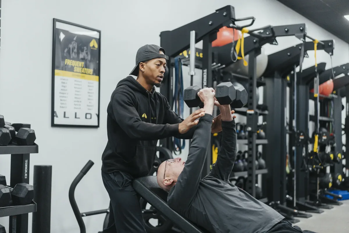 A coach guides an older member's dumbbell chest press from behind the bench