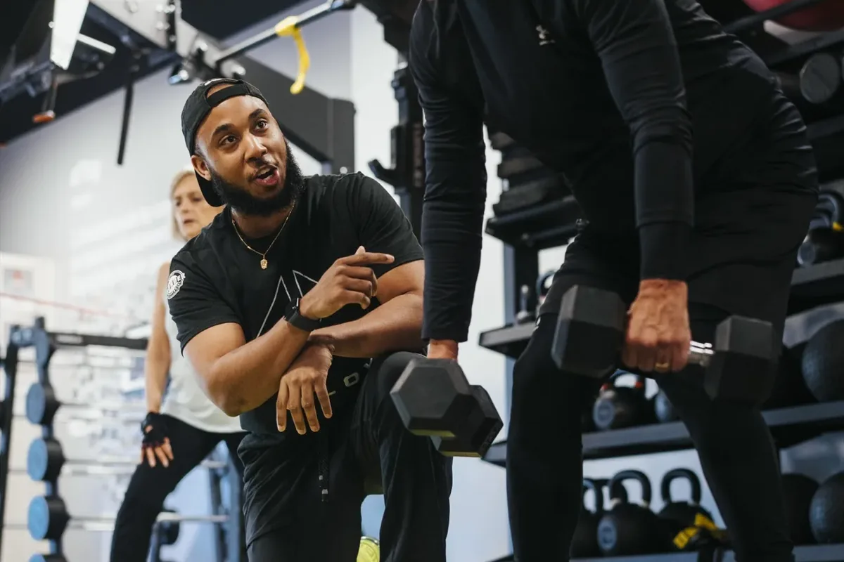 A coach kneels to cue an older member through bent-over dumbbell rows