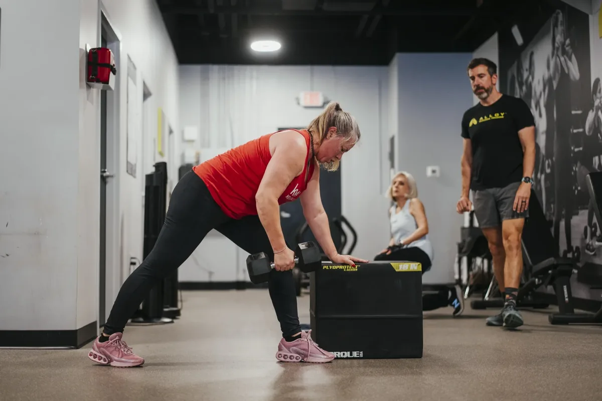 A member rows a dumbbell in a hinged position while a coach watches from a few feet away