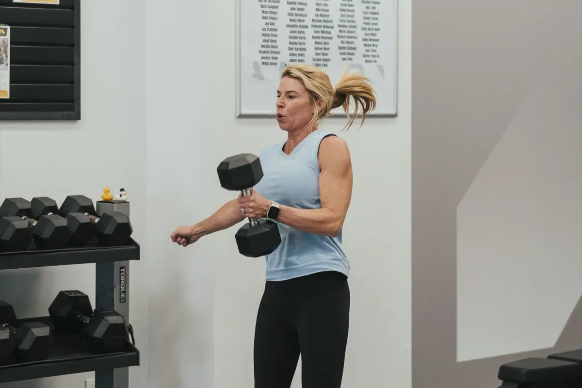 A woman powers through a standing dumbbell exercise beside the rack wall at an Alloy studio