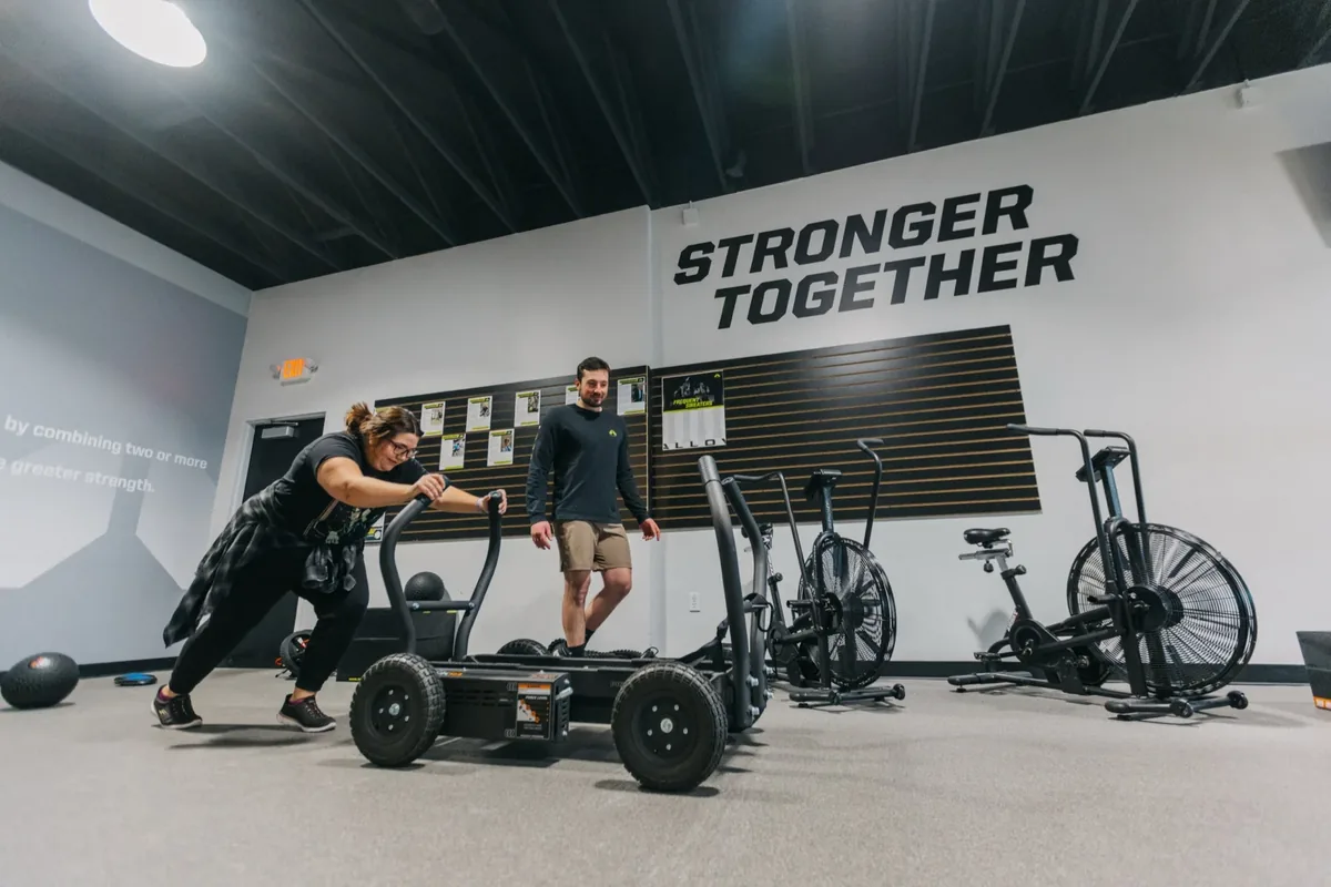 A coach looks on as a member pushes a weighted sled across the studio floor below the Stronger Together wall