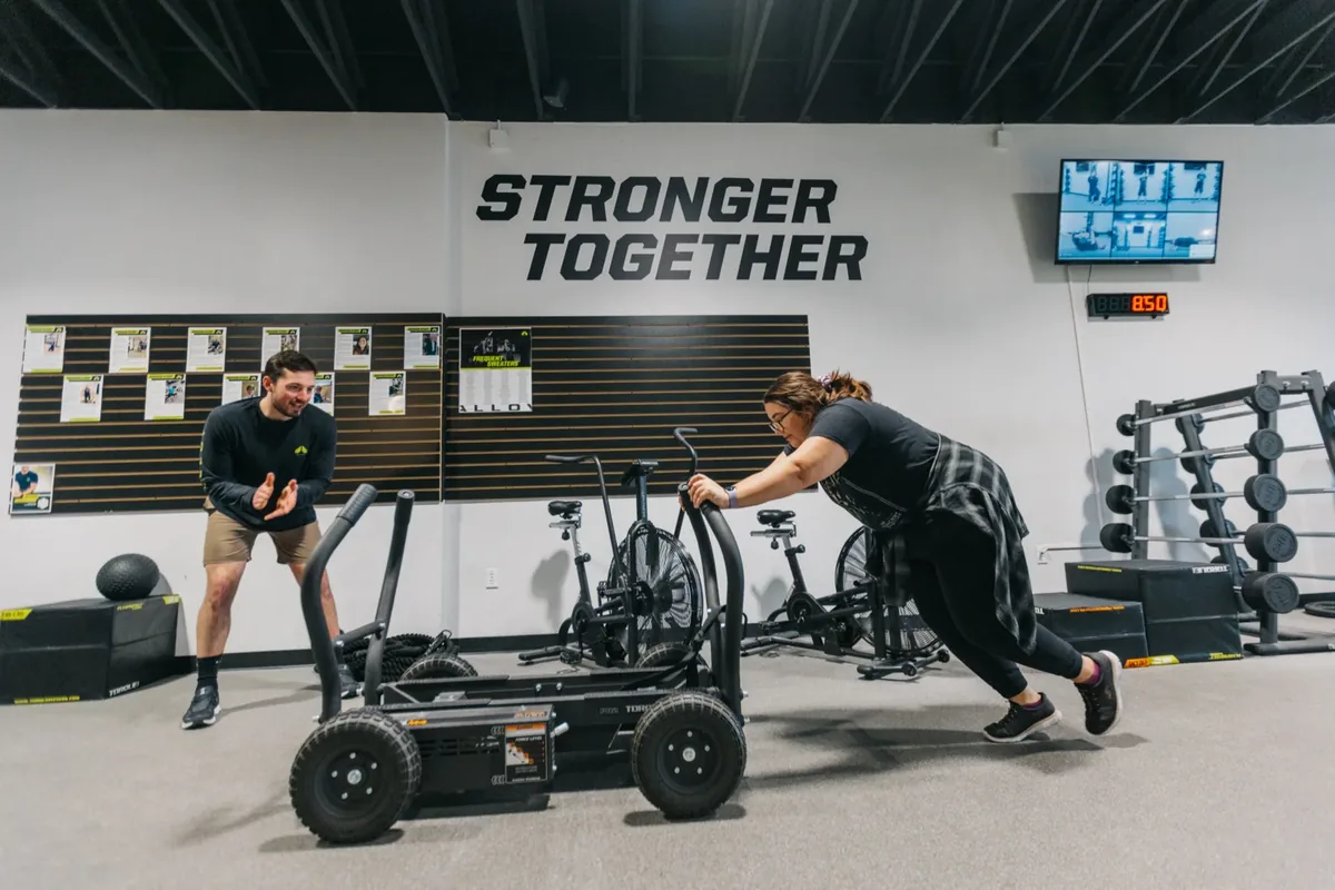A coach claps in encouragement as a member drives a weighted sled in front of the Stronger Together wall