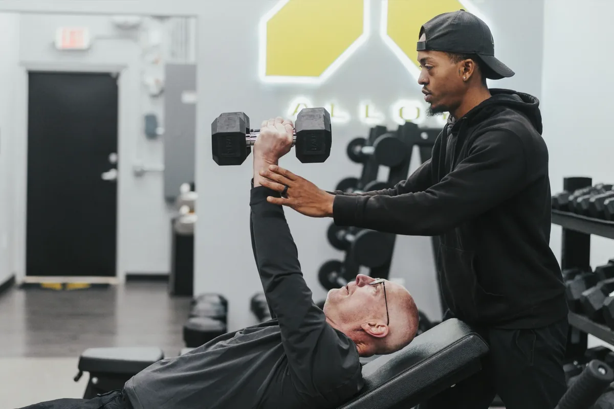 A coach spots an older member pressing dumbbells on an incline bench