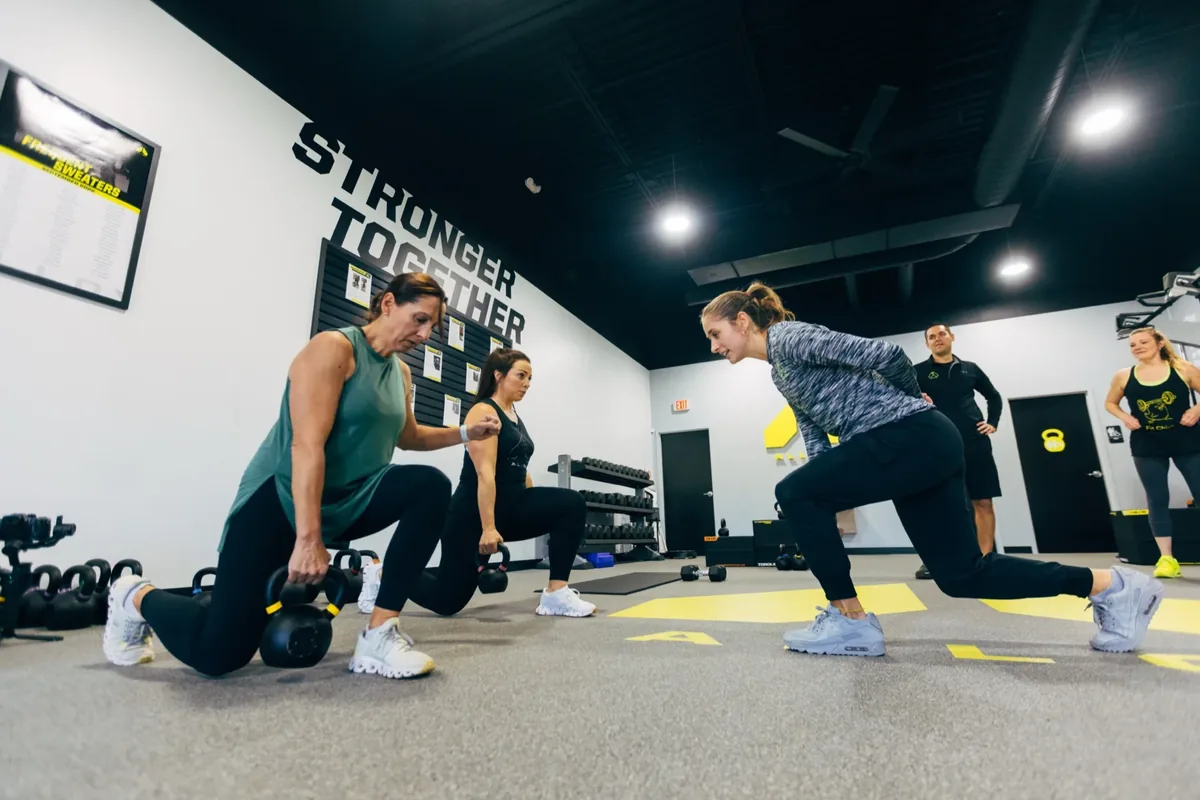 A coach leads three members through kettlebell lunges during a small group session at an Alloy studio
